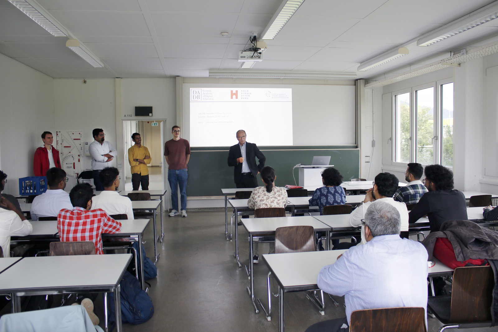 Ein Mann im Anzug steht vor sitzenden Teilnehmenden im Hintergrund ist auf eine Leinwand eine Präsentation zu erkennen 