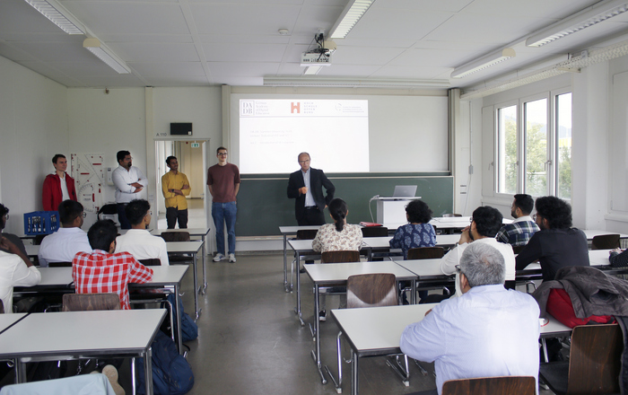 Ein Mann im Anzug steht vor sitzenden Teilnehmenden im Hintergrund ist auf eine Leinwand eine Präsentation zu erkennen 