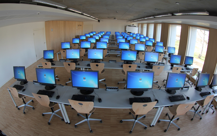 Ein menschenleerer Hörsaal mit ganz vielen eingeschalteten Computerbildschirmen.