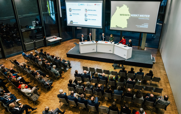 2025-10-27 Web-Meldung Hochschulen im Dialog Mehrere Menschen stehen auf einer Bühne im Halbkreis an Rednerpulten hinter ihne zwei große Bidlschirme mit Präsentationen und vor ihnen sitzende Zuhörende