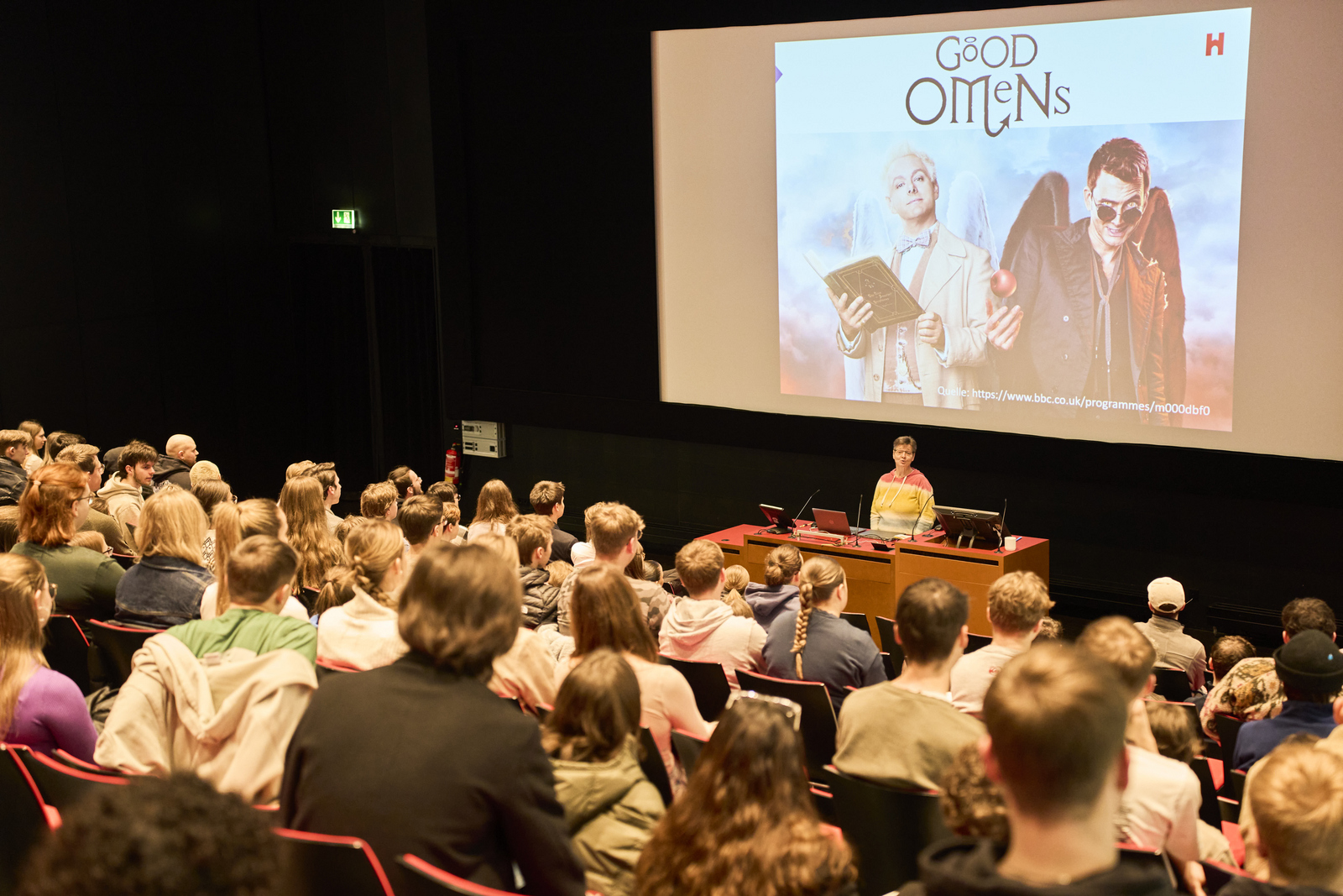 Eine Frau steht vor sitzenden jungen Menschen in einem Hörsaal hinter ihr auf eine Leinwand ist das Titelbild der Serie Good Omens zu sehen