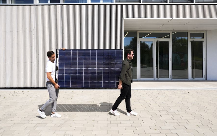 Zwei junge Männer tragen ein Photovoltaik-Modul über einen Platz vor einem Gebäude 