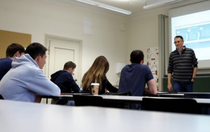 Eine Referent steht vor einer Gurppe Studierenden im Hintergrund ein eingeschaltetet Bildschirm.