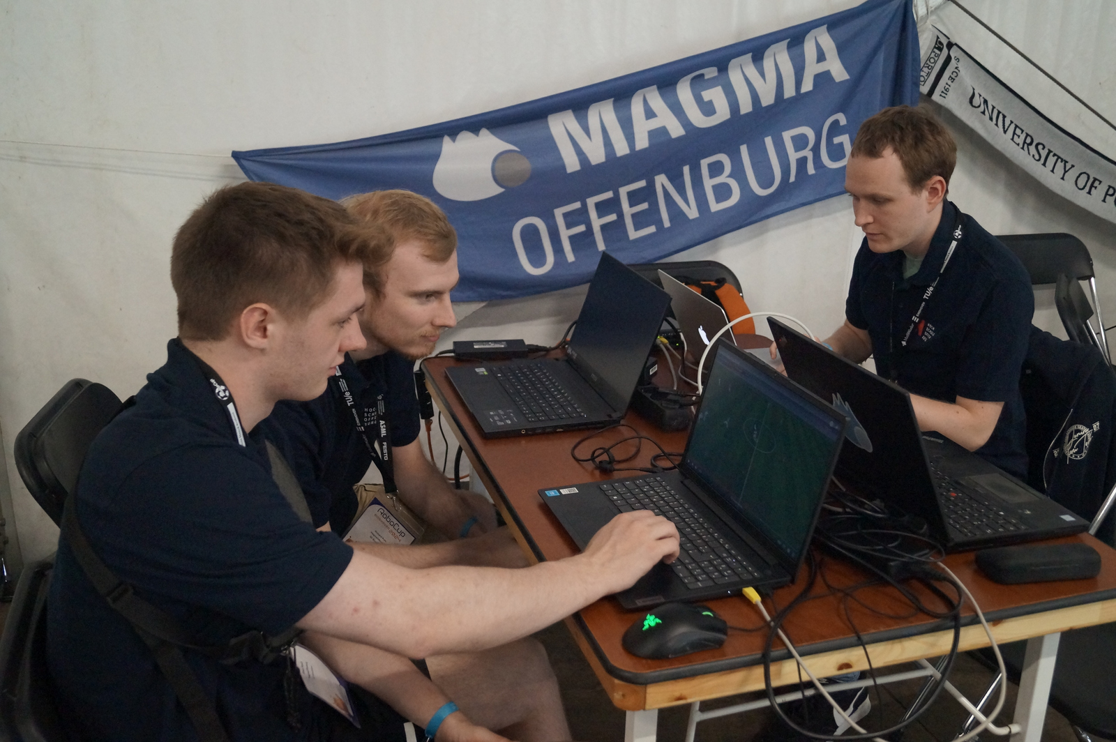 Drei junge Männer sitzen um einen Tisch mit mehreren Laptops im Hintergrudn hängt die Team-Fahne