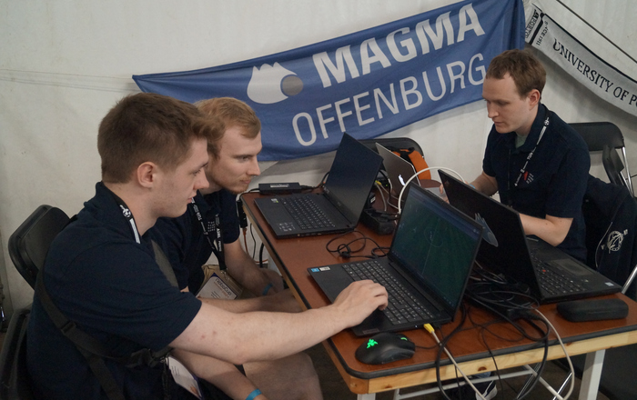 Drei junge Männer sitzen um einen Tisch mit mehreren Laptops im Hintergrudn hängt die Team-Fahne