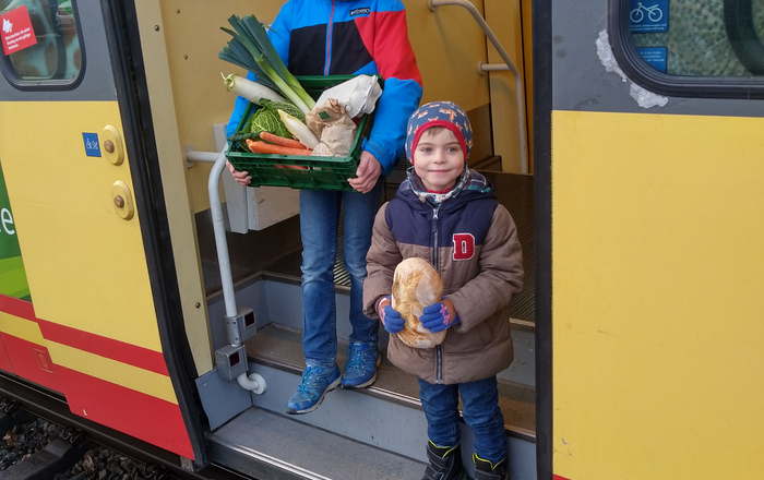Zwei Jungen mit eine Kiste Gemüse beziehungsweise Brot in der Hand steigen aus einer Stadtbahn