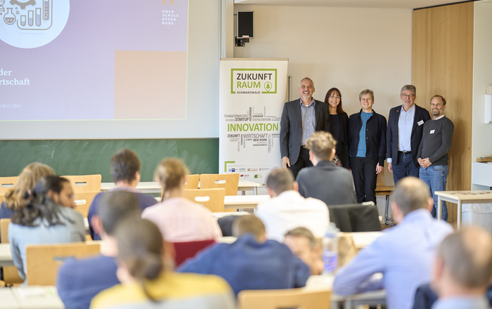 2024-12-02 PM Unternehmen Zukunft mit Institute for Circular Bioeconomy Blick über sitzenden Leute hin zu mehreren Personen, die rechts neben einem Roll-Up stehen