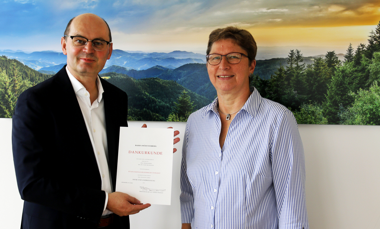 Ein Mann im Anzug überreicht einer Frau in Bluse eine Urkunde, die beiden stehen vor einem Schwarzwaldfoto