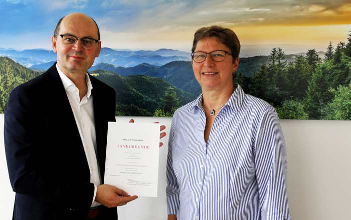 Ein Mann im Anzug überreicht einer Frau in Bluse eine Urkunde, die beiden stehen vor einem Schwarzwaldfoto