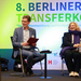 Ein Mann und eine Frau sitzen auf einer Bühne hinter in steht 8. Berliner Transferkonferenz 