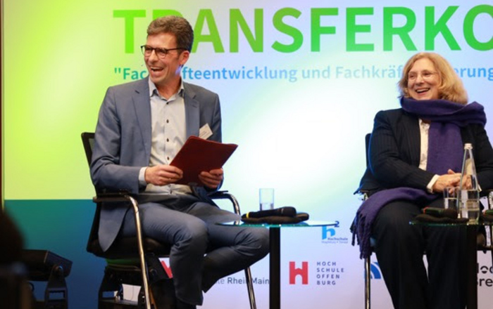 Ein Mann und eine Frau sitzen auf einer Bühne hinter in steht 8. Berliner Transferkonferenz 