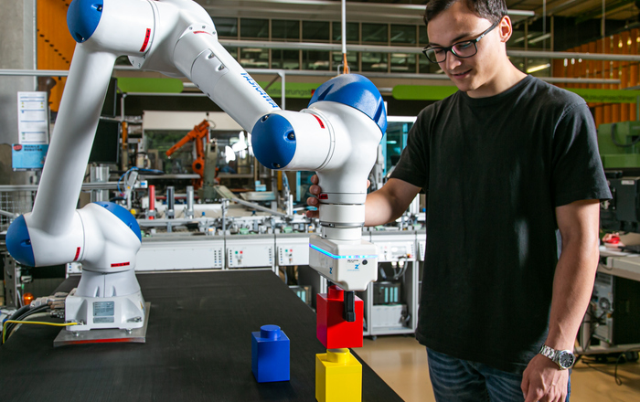 Ein junger Mann mit schwarzem T-Shirt und schwarzer Hose steht in einem Labor an einem Roboterarm der bunte Bauklötze stapelt