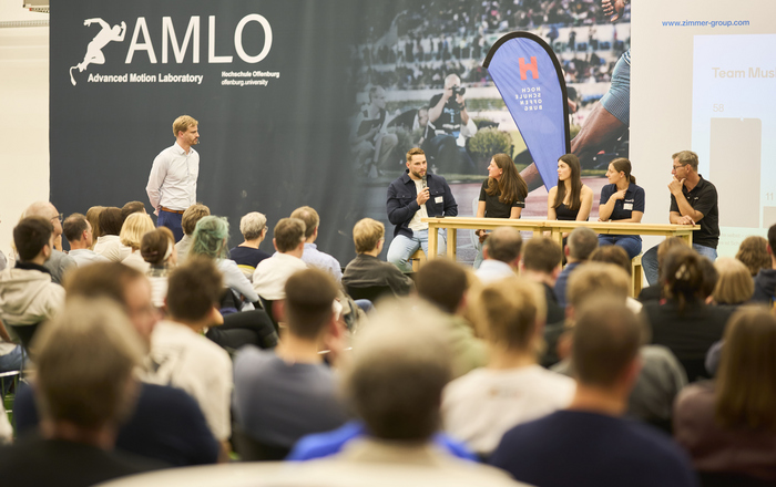 2025-10-13 PM 1. Industrie-Hochschulforum Biomechanik Podiumsdiskussion Blick über sitzende Menschen hinweg auf die Bühne wo vier Personen sitzen ein Mann steht links von ihnen