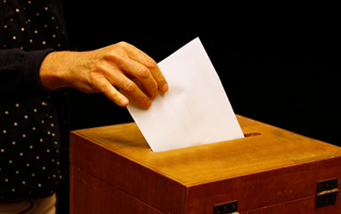 Eine weibliche Hand steckt einen Wahlzettel in eine hölzerne Wahlurne.