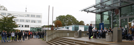 Das Hochschulorchester spielt vor Gebäude D und einige Zuhörer stehen auf dem Campus.