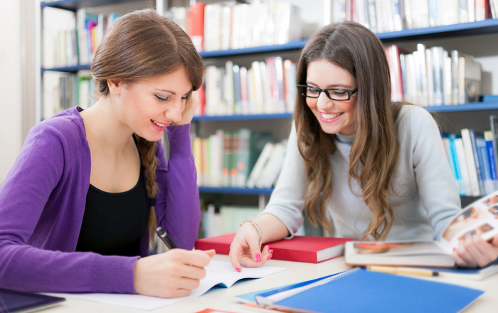 Zwei Studentinnen sitzen vor einem Bücherregal an einem Tisch und lernen.