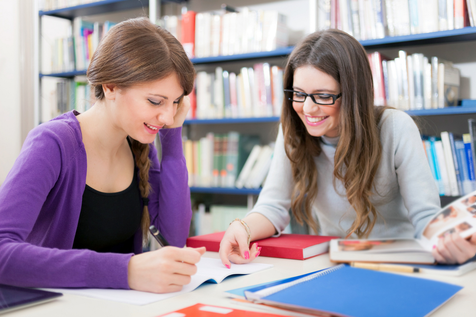 Zwei Studentinnen sitzen vor einem Bücherregal an einem Tisch und lernen.