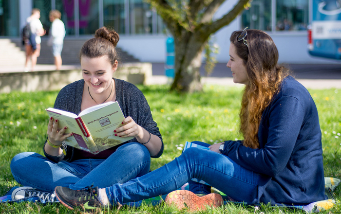 Zwei Studentinnen sitzen auf der Campus-Wiese, eine liest in einem Buch.