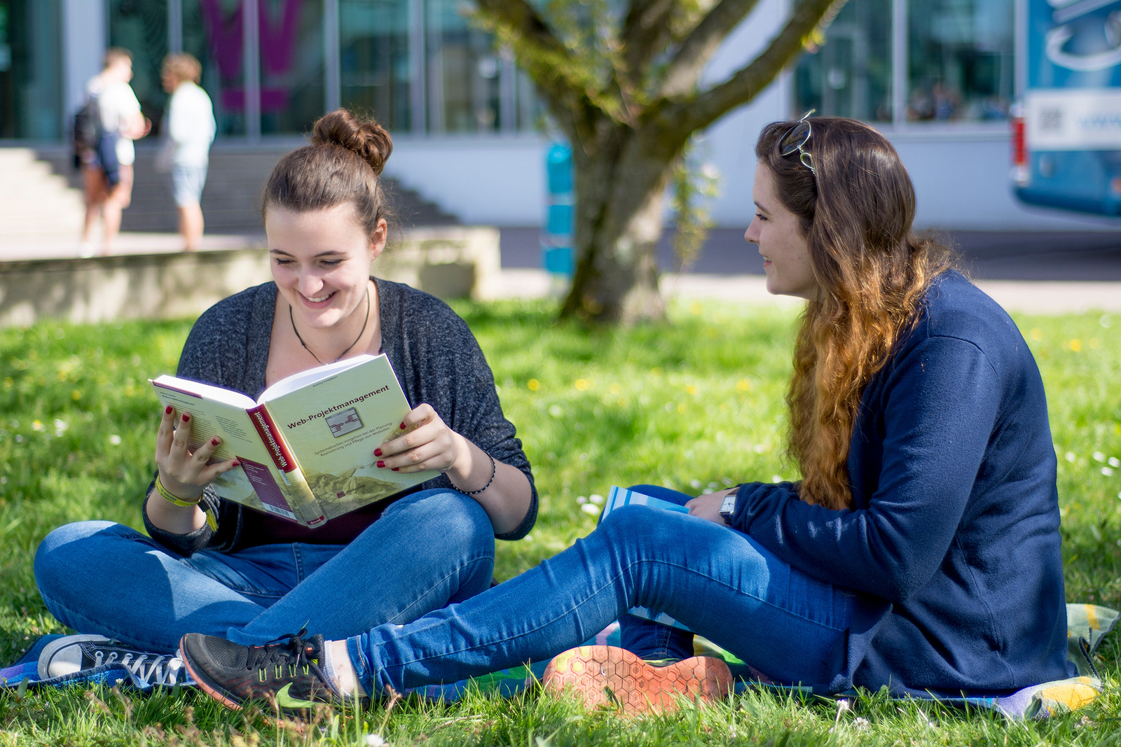 Zwei Studentinnen sitzen auf der Campus-Wiese, eine liest in einem Buch.