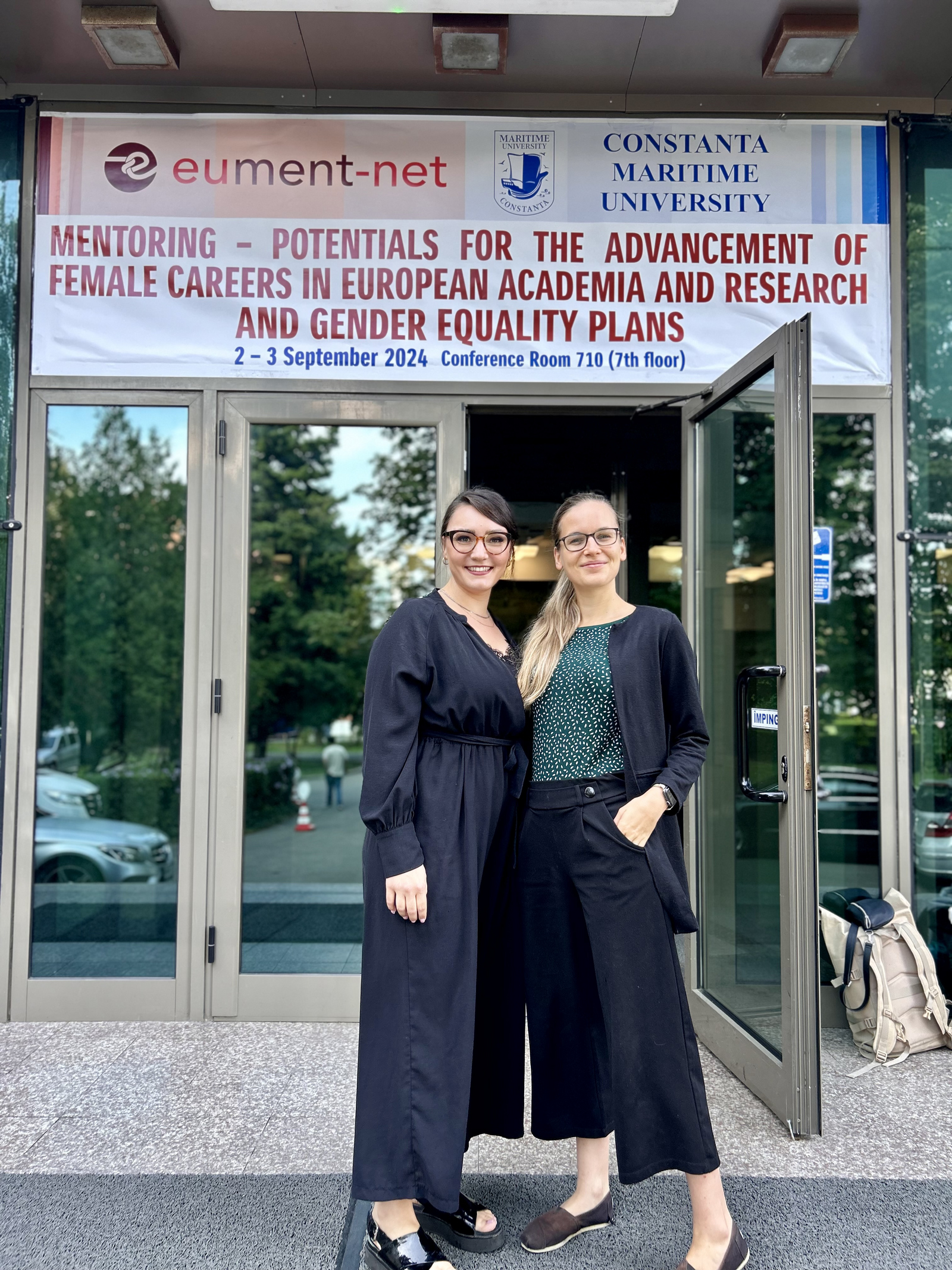 Zwei Frauen stehen vor einer geöffneten Glastür über der ein weißes Banner mit dem Konferenznamen und -datum in rot und blau hängt.