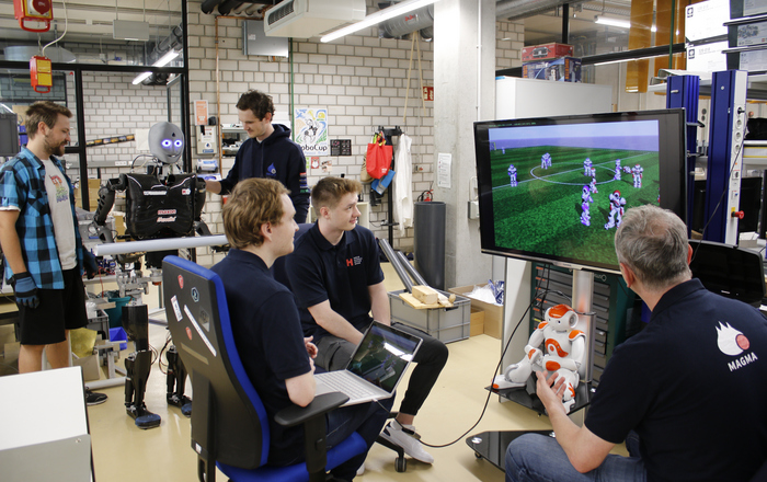 Team Magma sitzt vor einem Bildschirm auf dem ein feld mit simulierten Robotern zu sehen ist, im Hintergrund steht Sweaty und stemmt beobachtet von zwei Teammitgliedern Gewichte