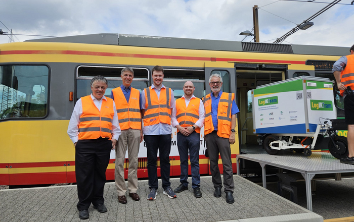 Fün Männer unterschiedlichen Alters in orangefarbenen Warnwesten stehen vor einem Straßenbahnwagen in den ein Lastenradanhänger hinfährt