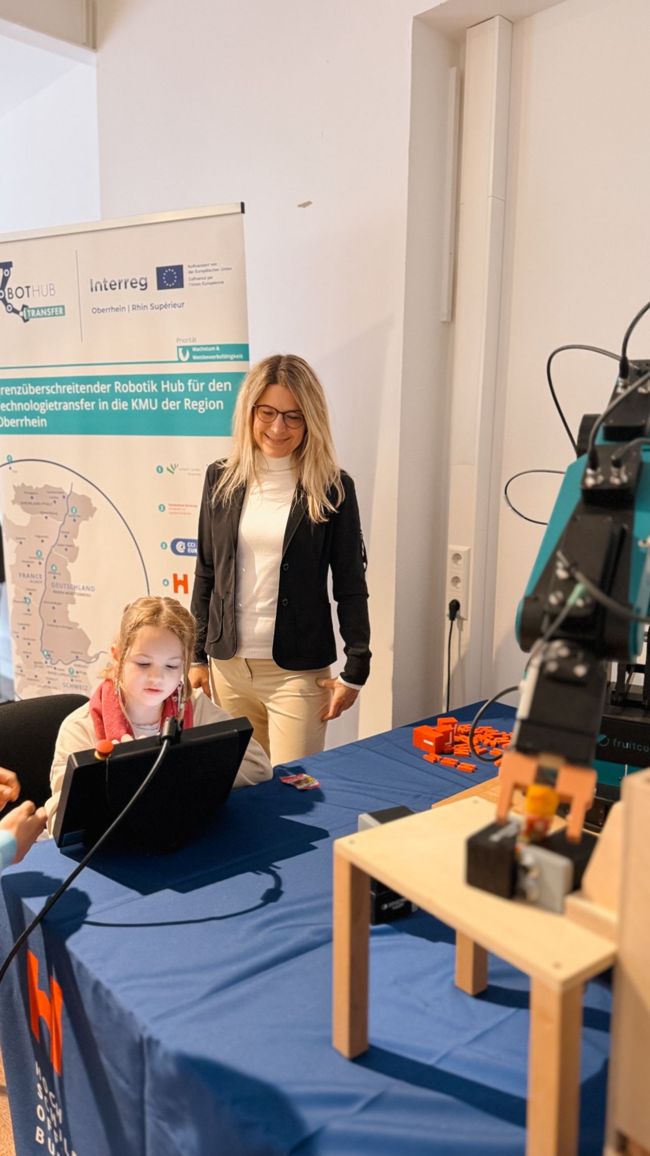 A woman stands behind a young girl, who is learning to control a robot with a table