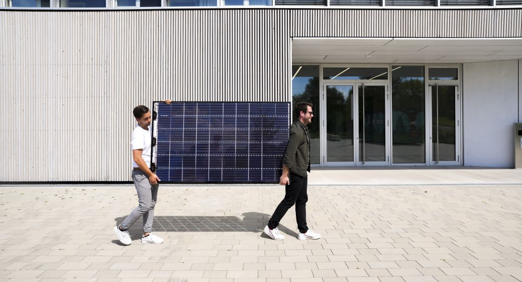 Zwei junge Männer tragen ein Photovoltaik-Modul über einen Platz vor einem Gebäude 