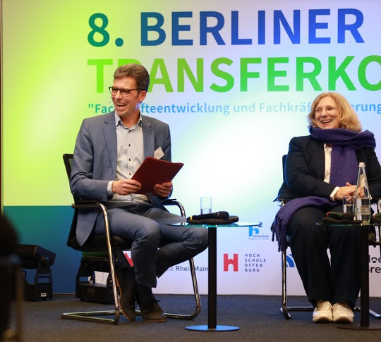 Ein Mann und eine Frau sitzen auf einer Bühne hinter in steht 8. Berliner Transferkonferenz 